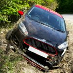 A black car, seemingly a Ford, crashed and damaged in a roadside ditch with surrounding greenery.