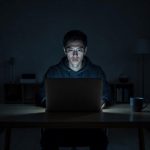 A young man at a laptop late at night, face lit by screen glow, expression distant
